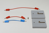 Zwei graue Batteriefächer und drei Anschlusskabel in Rot und Blau liegen auf hellem Grund. Ergänzt wird das Set durch eine kleine Glühbirne für physikalische Experimente.